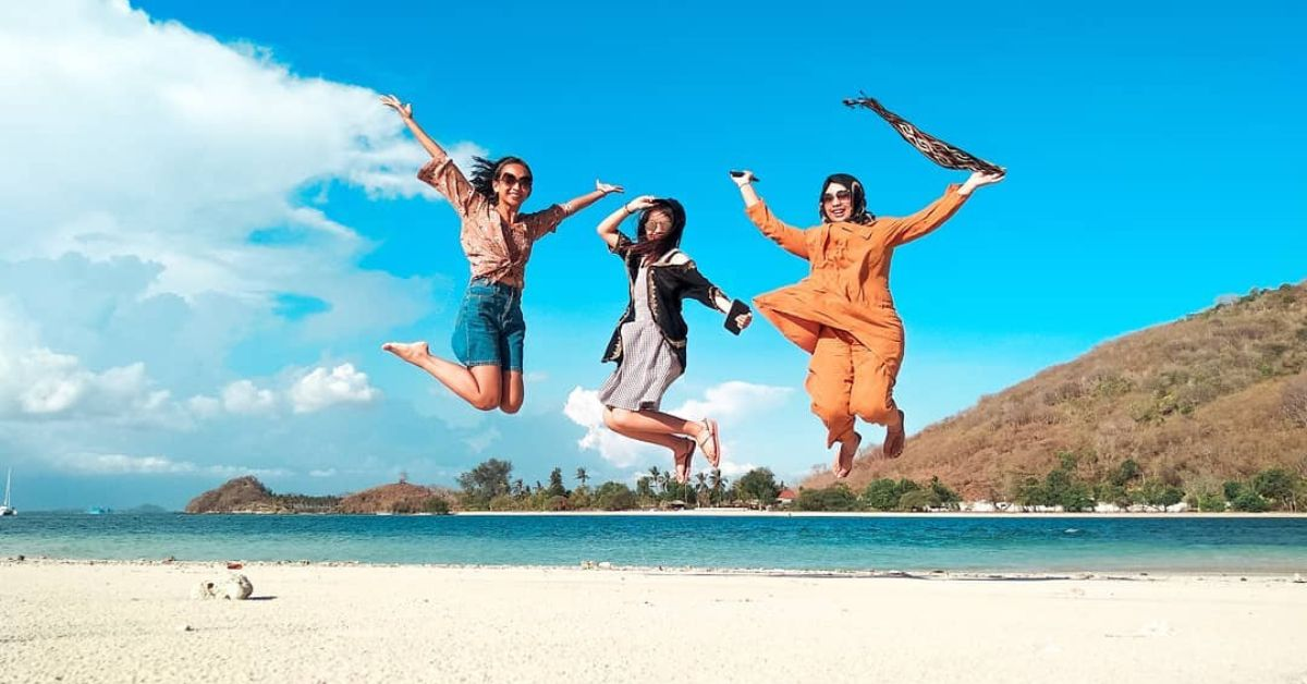 Tiga wisatawan wanita melompat gembira di pantai indah Lombok dengan latar laut biru dan bukit hijau