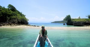 Wisatawan berpose di ujung perahu dengan latar pantai tersembunyi dan air laut jernih saat mengikuti paket trip Lombok 3 hari 2 malam Opsi C
