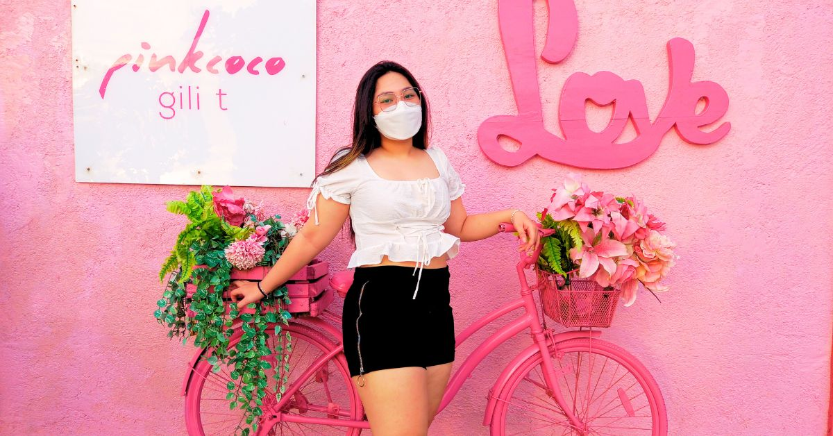 Wisatawan berpose di depan dinding pink dengan sepeda dan bunga di PinkCoco Gili Trawangan saat tur Explore Lombok 4 Hari 3 Malam