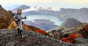 Pendaki berdiri di jalur puncak Gunung Rinjani dengan latar Danau Segara Anak – bagian dari paket trekking Gunung Rinjani.