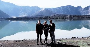 Tiga pendaki berpose di tepi Danau Segara Anak dengan latar Gunung Barujari – bagian dari paket trekking Gunung Rinjani.