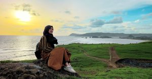 Seorang wanita duduk santai di atas bukit hijau berlatar belakang laut dan langit senja di Lombok.