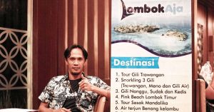 Seorang anggota tim Lombok Aja Tour Organizer duduk di samping banner destinasi wisata Lombok, memberikan jempol sebagai simbol layanan agen travel Lombok yang profesional.