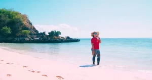 Seorang wisatawan berdiri di pasir berwarna pink di Pantai Tangsi, Lombok Timur dengan latar belakang laut biru yang tenang.